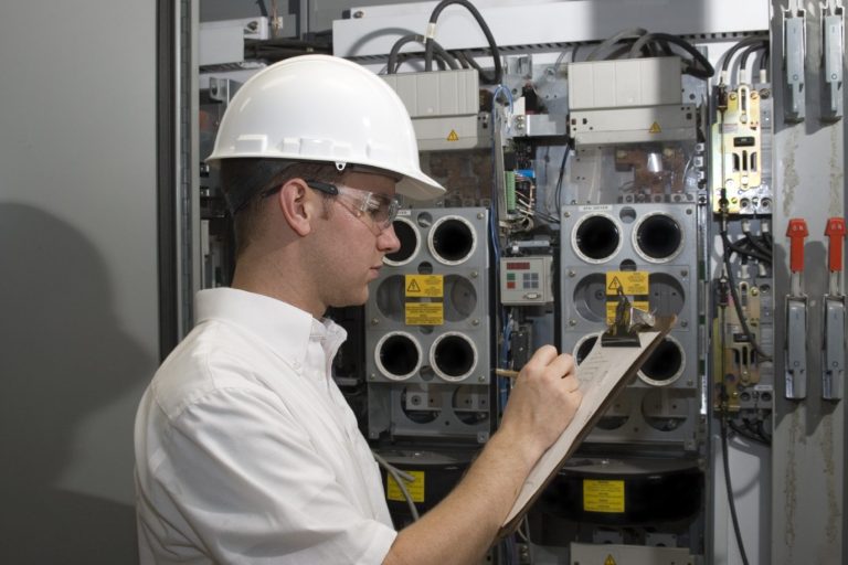 Technicien en casque et lunettes de sécurité, prenant des notes devant un panneau électrique.