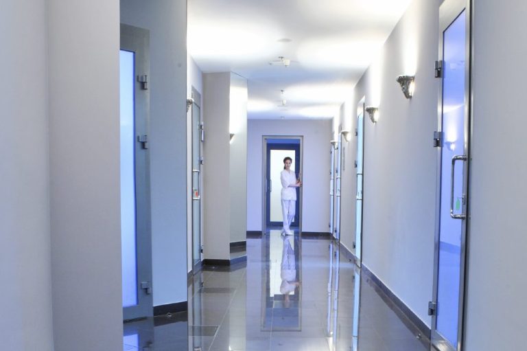 Couloir lumineux avec portes bleues et une personne en blouse blanche au fond.